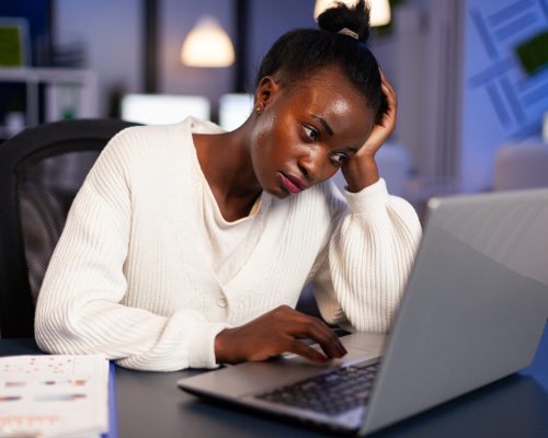 Exhausted african freelancer resting head on hand in front of laptop working overtime in start up company office. Stressed employee using modern technology doing overtime for deadline of project.