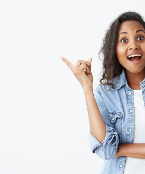 Human face expressions, emotions and feelings. Astonished and surprised young Afro-american female in light-blue denim shirt pointing at blank studio wall, surprised with sale prices, keeping her mouth wide open, demonstrating her white teeth.