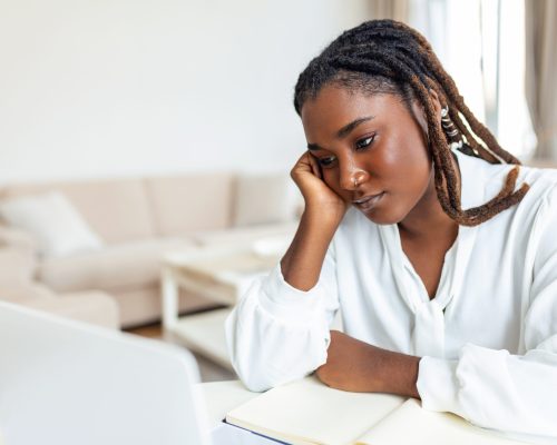 Serious frowning African American ethnicity woman sit at workplace desk looks at laptop screen read e-mail feels concerned. Bored unmotivated tired employee, problems difficulties with app